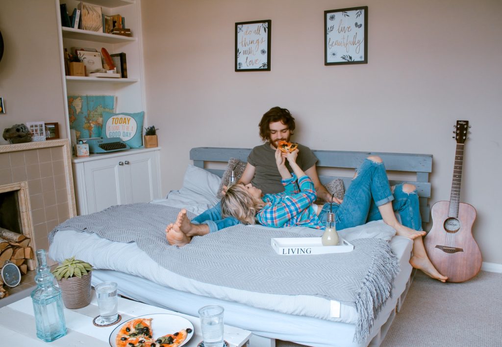 A man and woman sit in bed eating pizza.