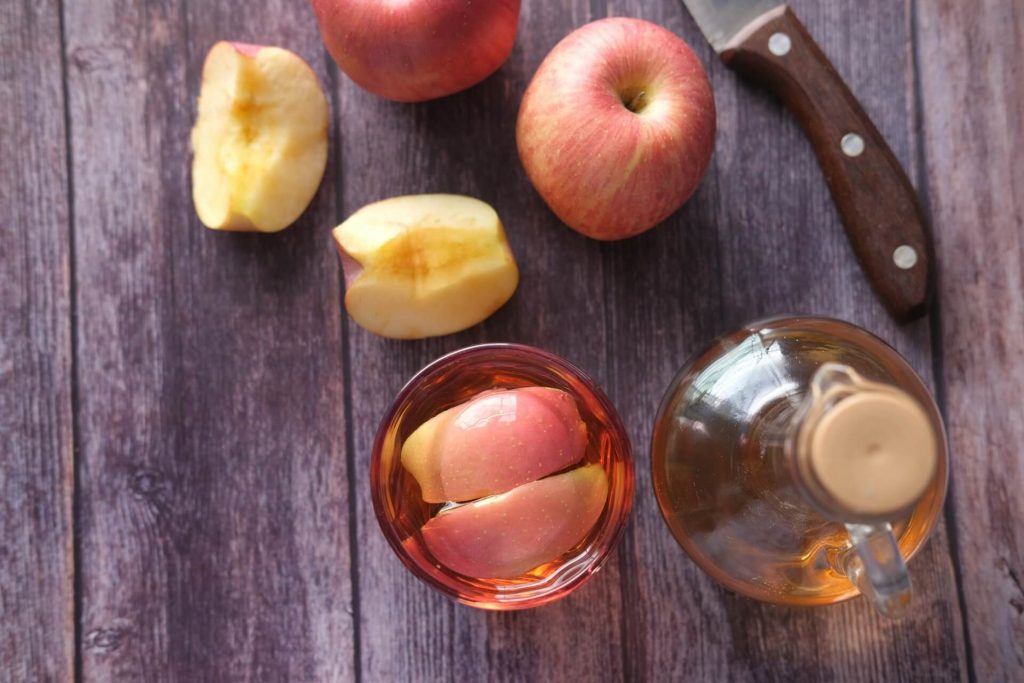 A glass of apple cider vinegar next to a bottle of apple cider vinegar and a knife with apples next to it