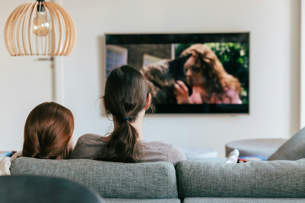 A woman and a girl sit on a couch watching TV.