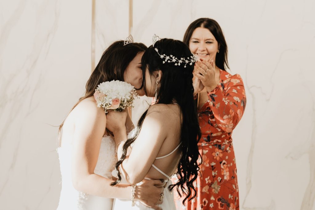 Two brides share a first kiss while the wedding officiant claps in background.