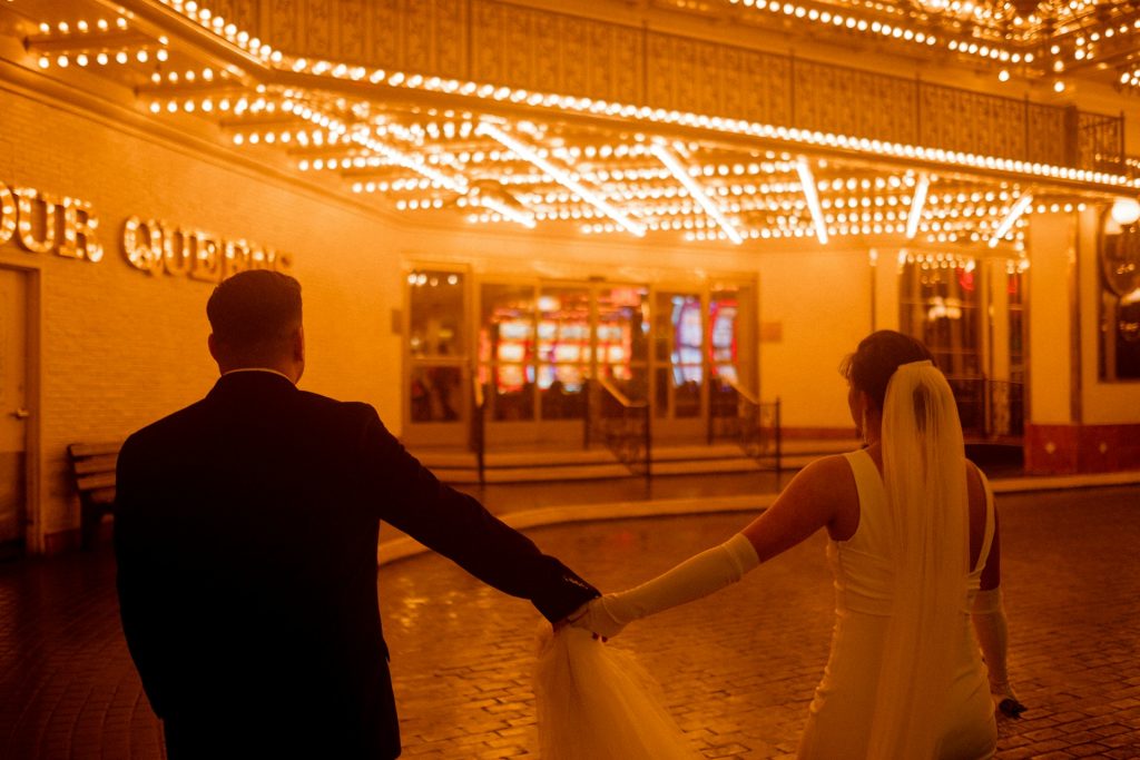 A bride and groom walk hand in hand toward a hotel door that waits under an array of orange-gold light bulbs that make the drive-up front door area glow.
