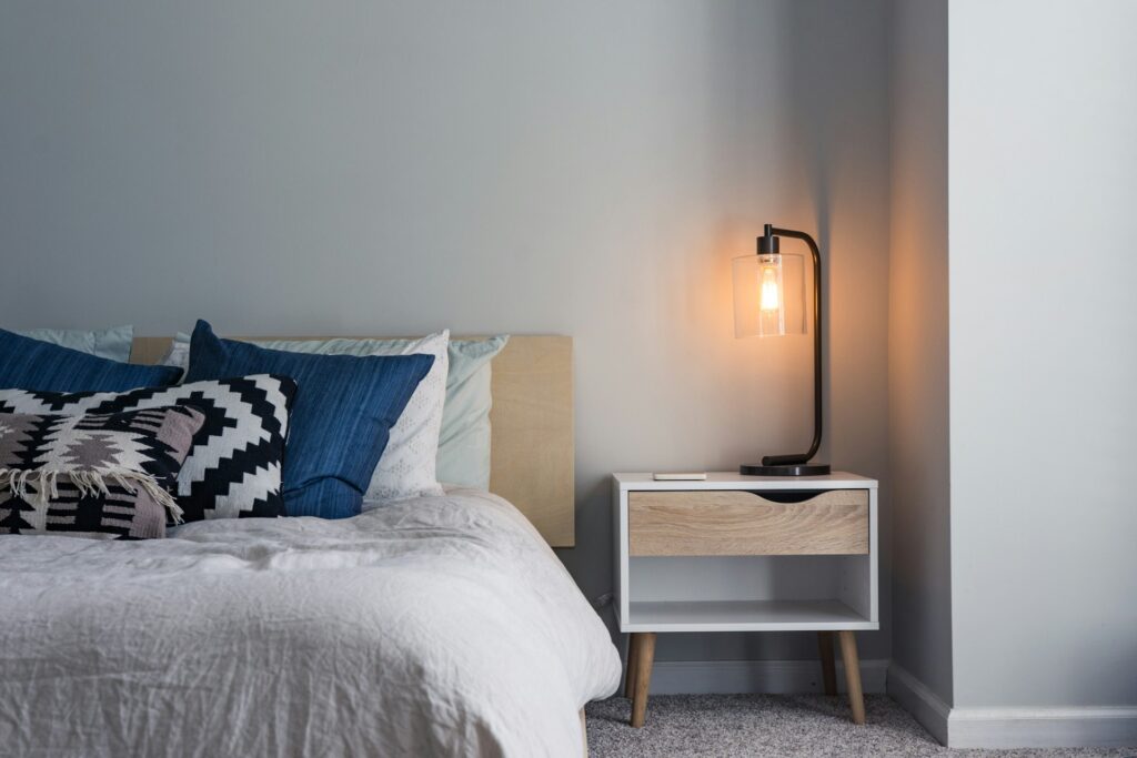 A bed features a white comforter, blue pillows and chevron throw pillows. A white night stand is next to the bed, holding a black metal lamp with an exposed bulb.