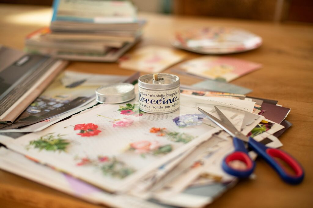 A cozy crafting scene with colorful papers featuring floral designs, a small jar of glue, and red-handled scissors on a wooden desk. Books and sheets of papers are in the background.
