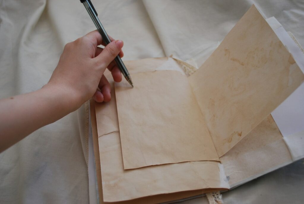A hand holding a pen hovers over an open notebook with aged, beige paper. The scene conveys a sense of nostalgia and contemplation.