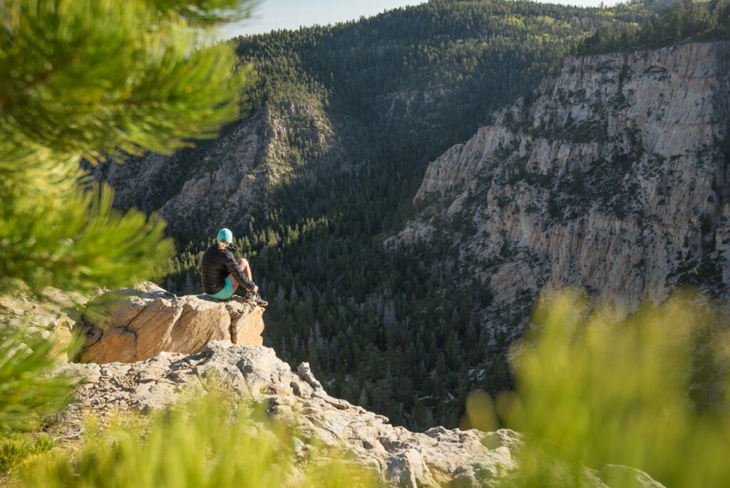 A woman wearing a black puffer jacket and a blue baseball cap sits on a rocky cliff overlooking other cliffs covered in trees as part of her nature therapy.