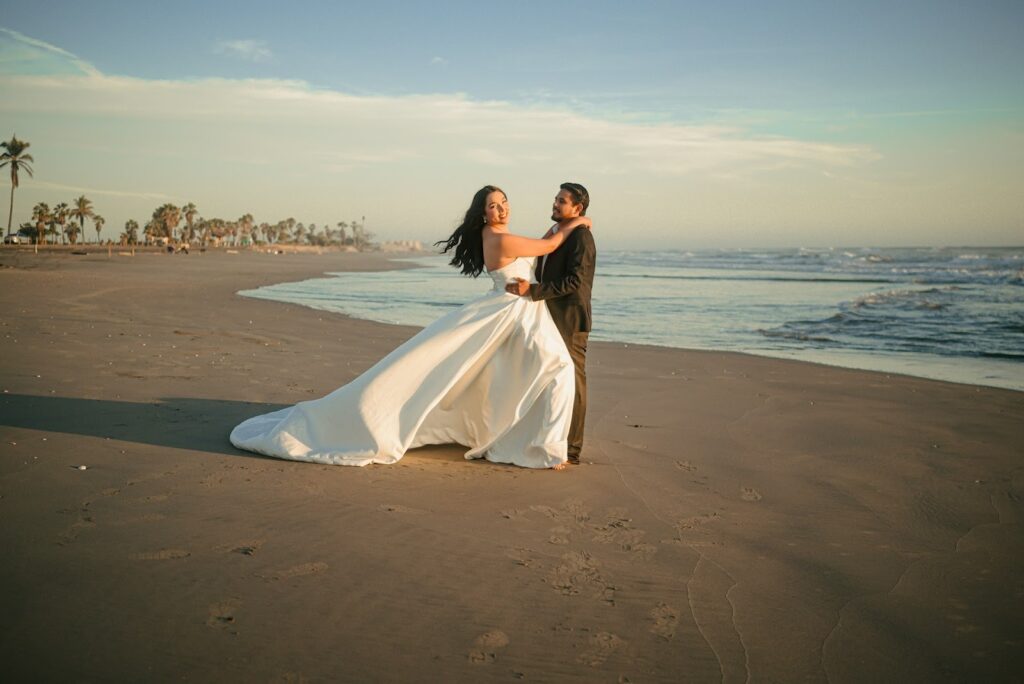 A couple dramatically poses by the beach, the bride wears a big, silky wedding gown while the groom looks at the bride fondly