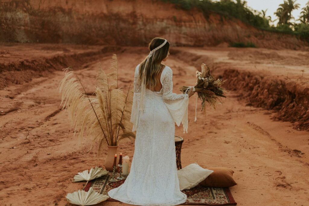 A woman in a white lace dress stands on a patterned rug in a desert setting, holding a bouquet. Pampas grass and candles create a rustic, serene atmosphere.