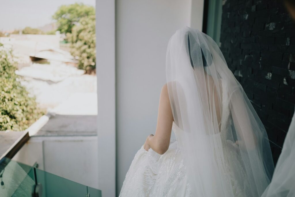 A bride is in a stunning white wedding dress and a long, translucent veil on a balcony, capturing a moment of celebration and beauty.