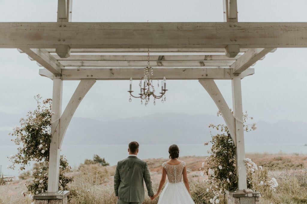A couple stands under a wooden pergola adorned with a chandelier and floral arrangements, facing a serene lake and mountain view, evoking romance and tranquility.