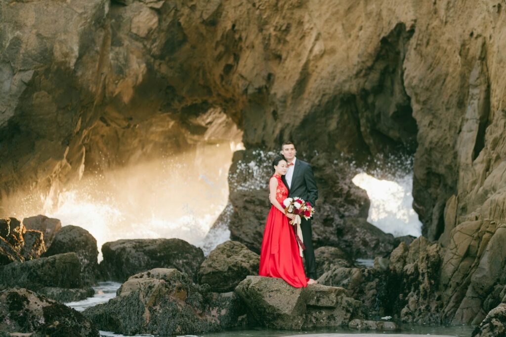 A couple at a rocky beach posing, the woman in a flowing red wedding dress holding a white and red bouquet, while the groom is in a tuxdeo
