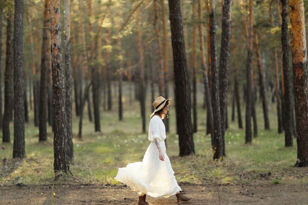 A woman wearing a white dress and straw hat walks gracefully through a serene forest, with sunlight filtering through the trees.