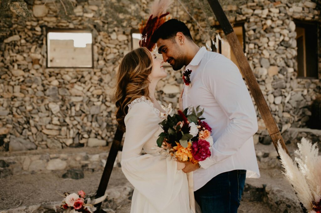 A couple shares a joyful moment in front of a rustic stone building. She holds a vibrant bouquet, while both gaze lovingly into each other's eyes.