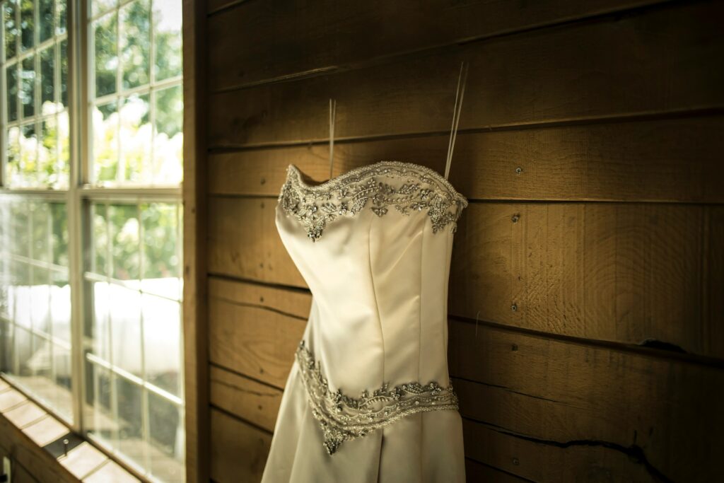 A silk wedding dress with silver embellishments elegantly hangs on a rustic wooden wall, showcasing its lustrous fabric and intricate design.