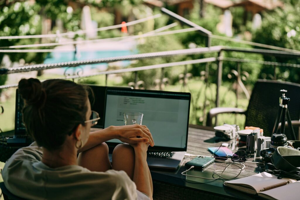 A person with a bun sits outdoors at a table with a laptop, surrounded by gadgets and a notebook, looking relaxed. Lush greenery is in the background.