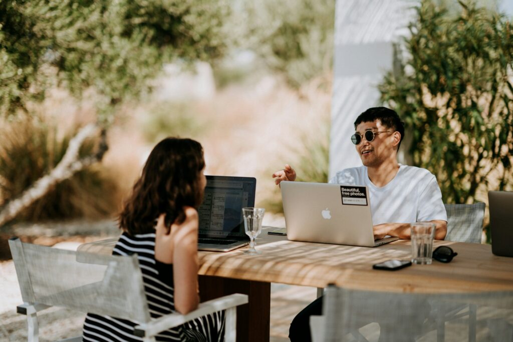 Two people sit outdoors at a wooden table with laptops. They're engaged in a lively discussion, surrounded by greenery and bathed in sunlight.