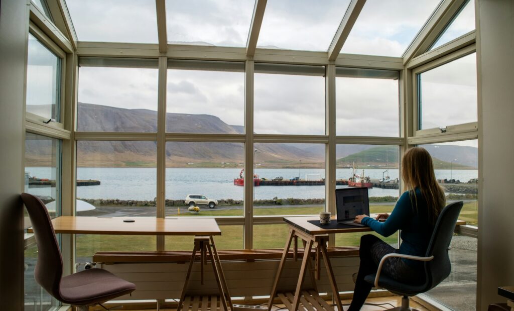 Woman working on a laptop in a bright room with large windows overlooking a serene lake and mountains. Interior exudes calm and focus.