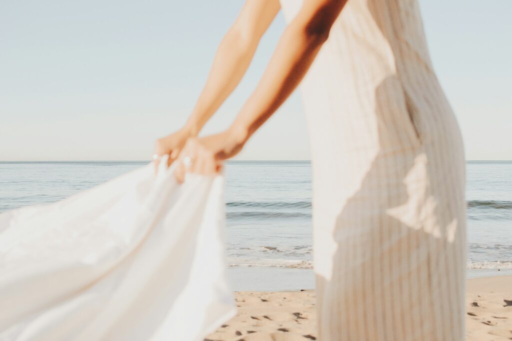A person in a light dress holds a white sheet on a sunlit beach. The sea is calm, creating a serene and airy atmosphere.