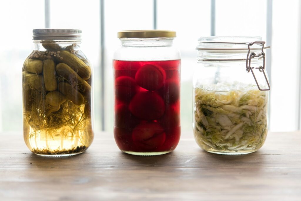 Three jars of preserved foods on a wooden surface: pickles, beets, and sauerkraut, with a bright, natural light streaming through a window behind them.