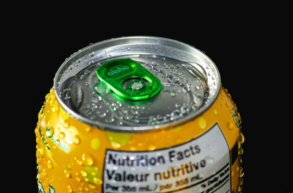 Close-up of a yellow soda can with a green tab, covered in water droplets against a black background. Part of the nutrition label is visible.