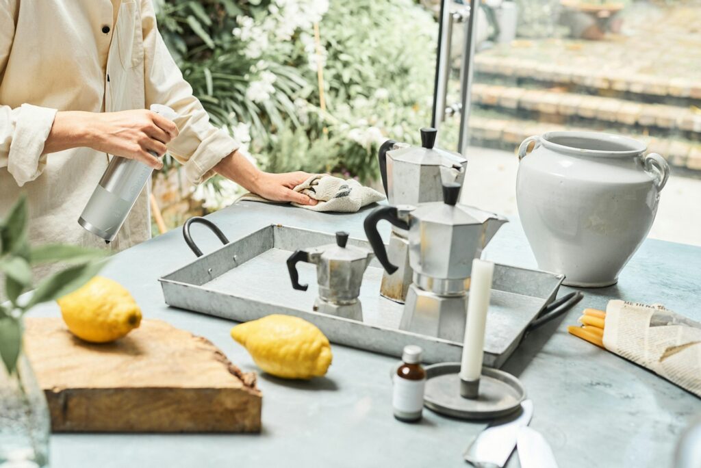 Person cleaning a tiled tabletop with a spray bottle and cloth. A metal tray with moka pots, lemons, a jar, and various items are placed nearby.