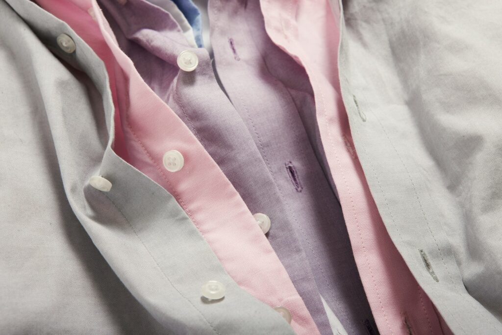 Close-up of three layered button-down shirts in light grey, pink, and lavender, showcasing texture and color contrast in soft lighting.