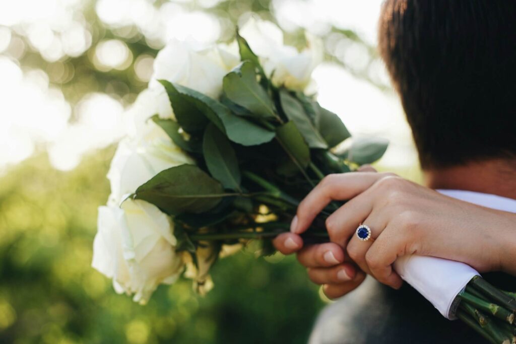 A bride wearing a sapphire ring surrounded by a diamond halo, holding a bouquet of flowers