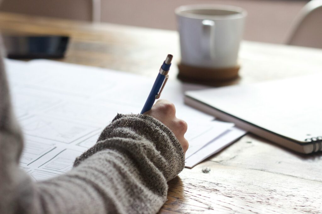 A person in a gray sweater writes on paper at a wooden table, with a notebook and a coffee cup nearby, conveying focus and productivity.