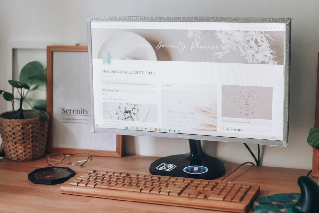 A wooden desk with a computer displaying a planner page. Nearby are framed art, a plant in a woven pot, and glasses, creating a serene workspace.