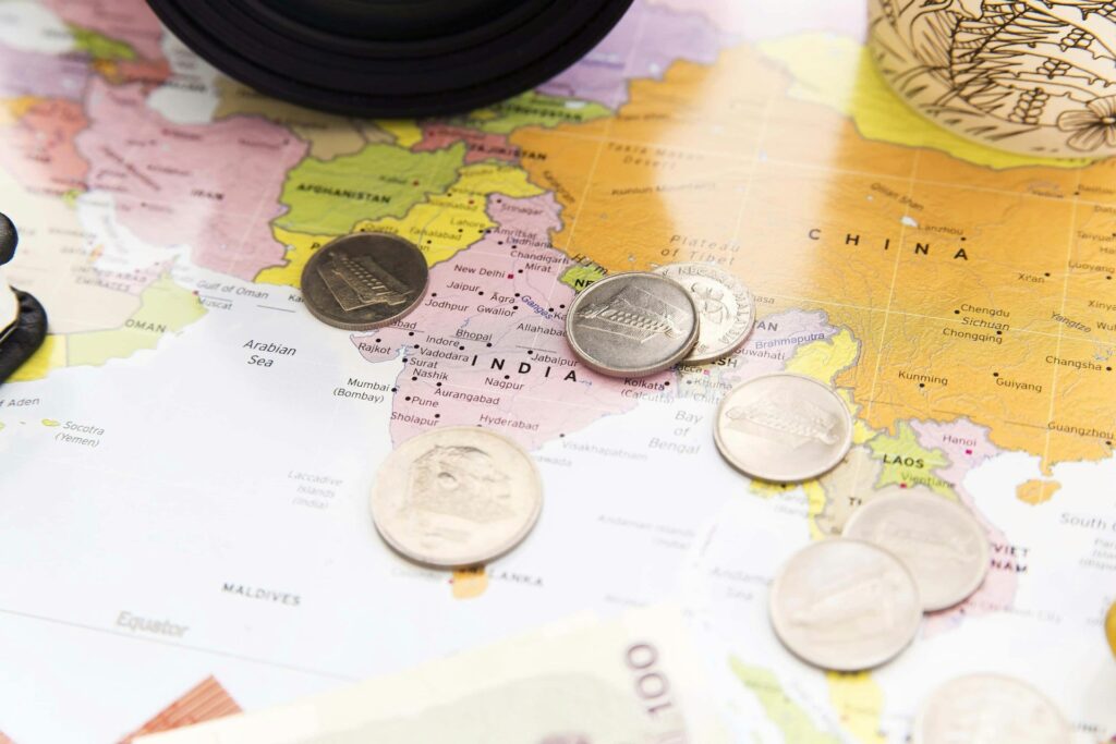 Coins scattered on a map of Asia, focusing on India. Items like a camera and cup edge frame the scene, suggesting travel planning.