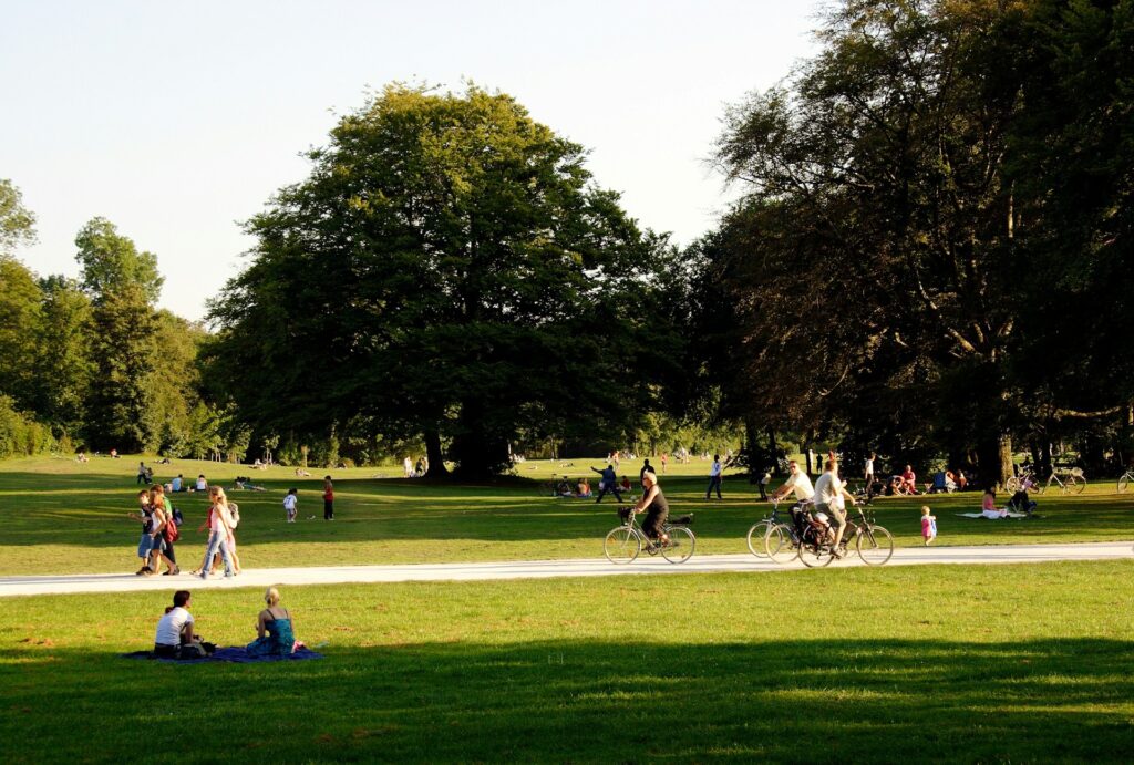 People enjoy a sunny park with green grass and large trees. Cyclists ride on a path, while others relax or stroll, creating a lively, peaceful scene.
