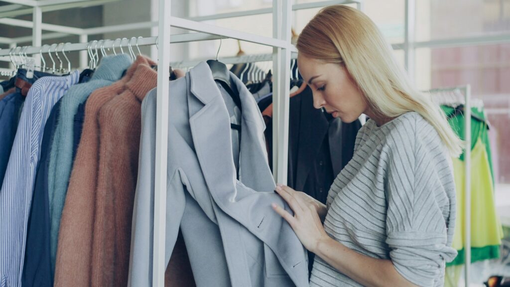 Woman in a gray sweater thoughtfully looks through clothes on a rack, examining a light gray blazer in a well-lit store. Shopping, decision-making.