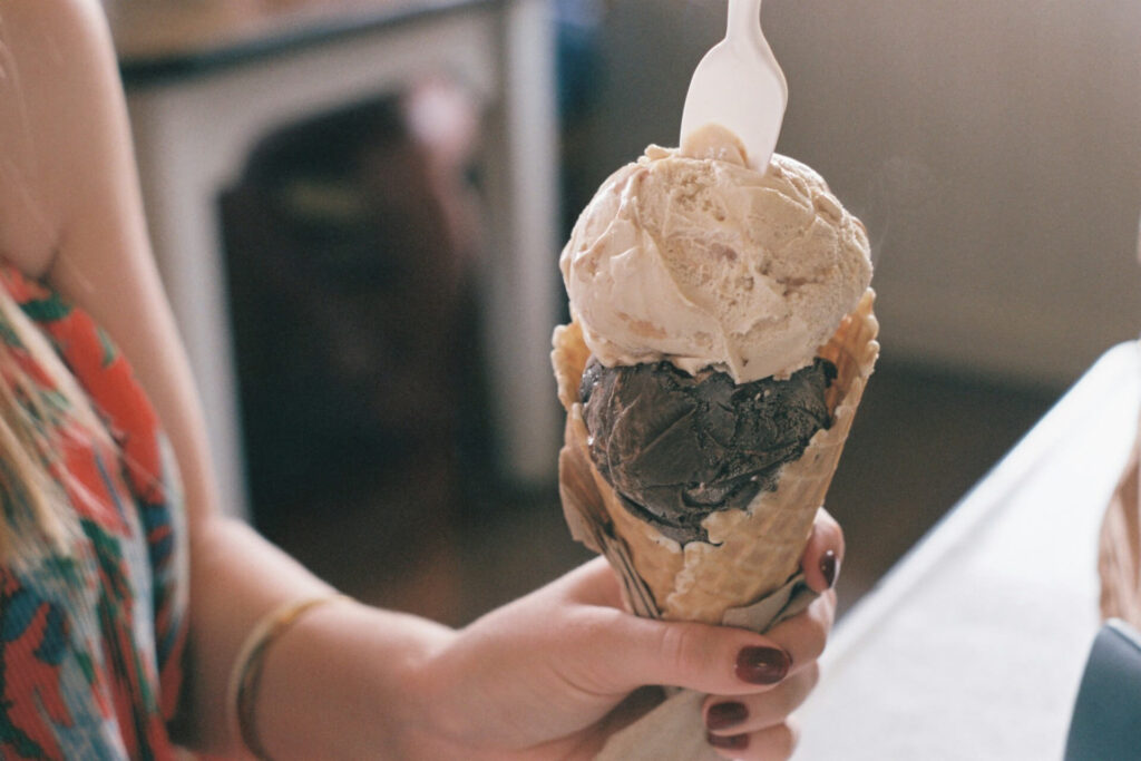 A person seated at a table, enjoying a two-scoop ice cream cone with a spoon