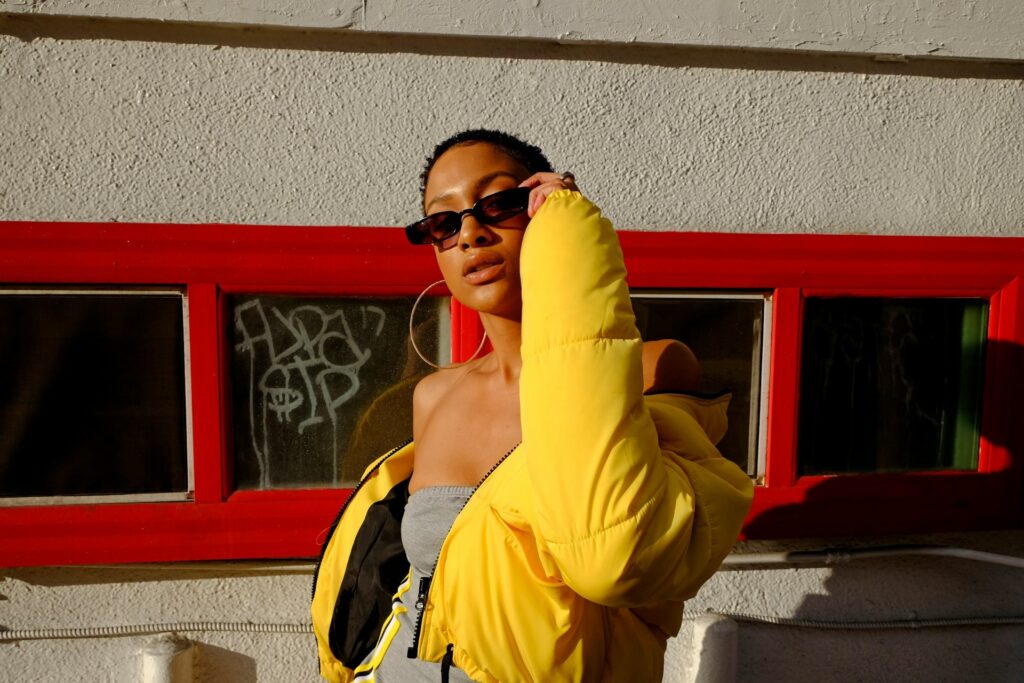 A woman in a yellow jacket and sunglasses poses outdoors, exuding a cheerful and stylish vibe.