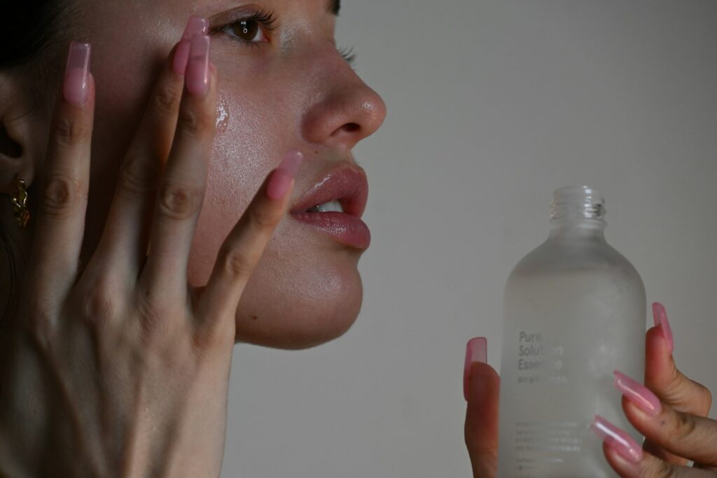 A woman with pink nails holds a frosted bottle of skincare, showcasing her manicured hands against a neutral background.