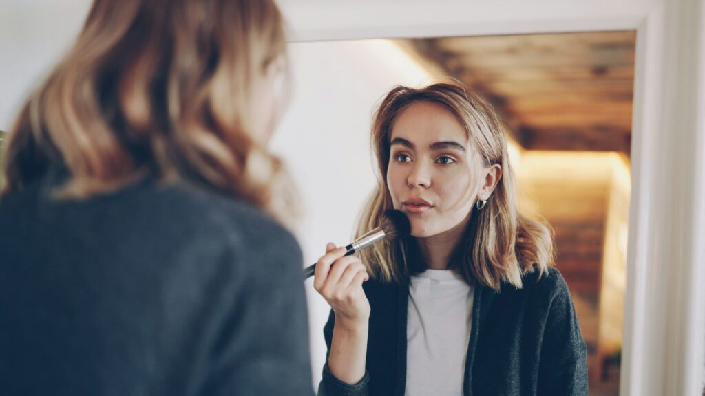 A woman with medium-length hair applies makeup with a brush while looking in a mirror. She appears focused, set in a warmly lit room with wooden decor.