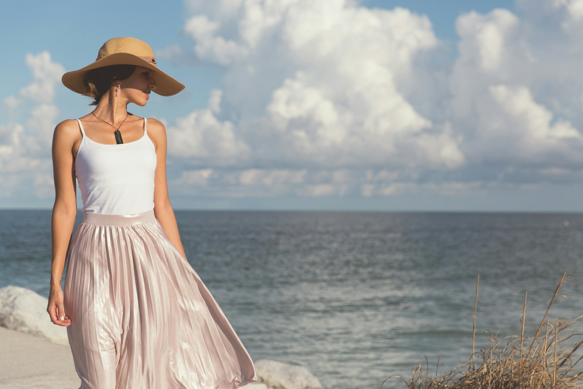 A woman in a stylish hat and skirt walks on the beach, enjoying the sun and the sound of the ocean waves.