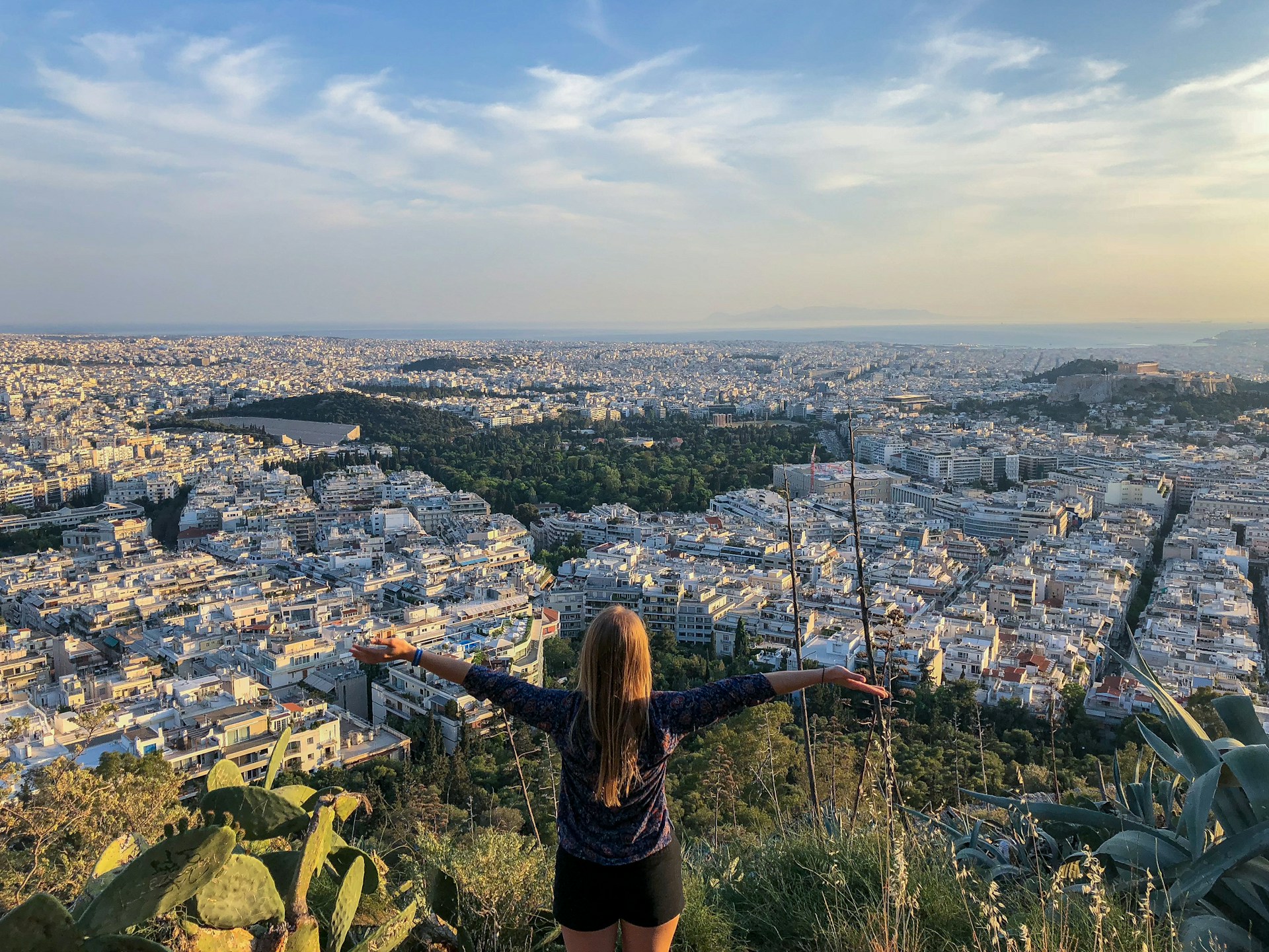A woman in a black shirt stands on a hill, gazing over a city with a panoramic view of the skyline.