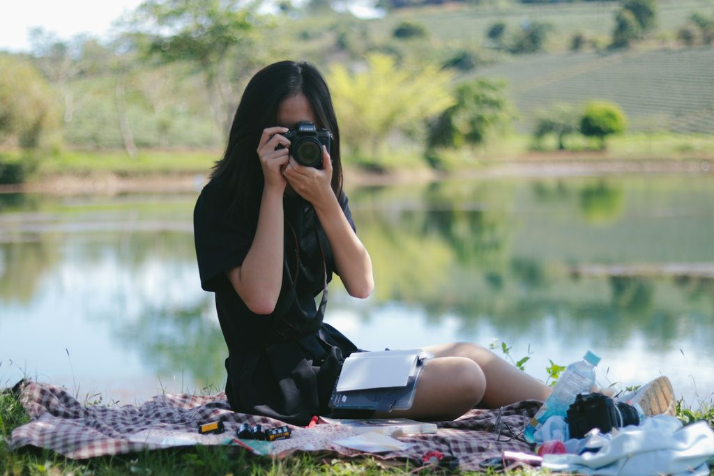 A woman sits on the grass, capturing a photo with her camera, surrounded by greenery.