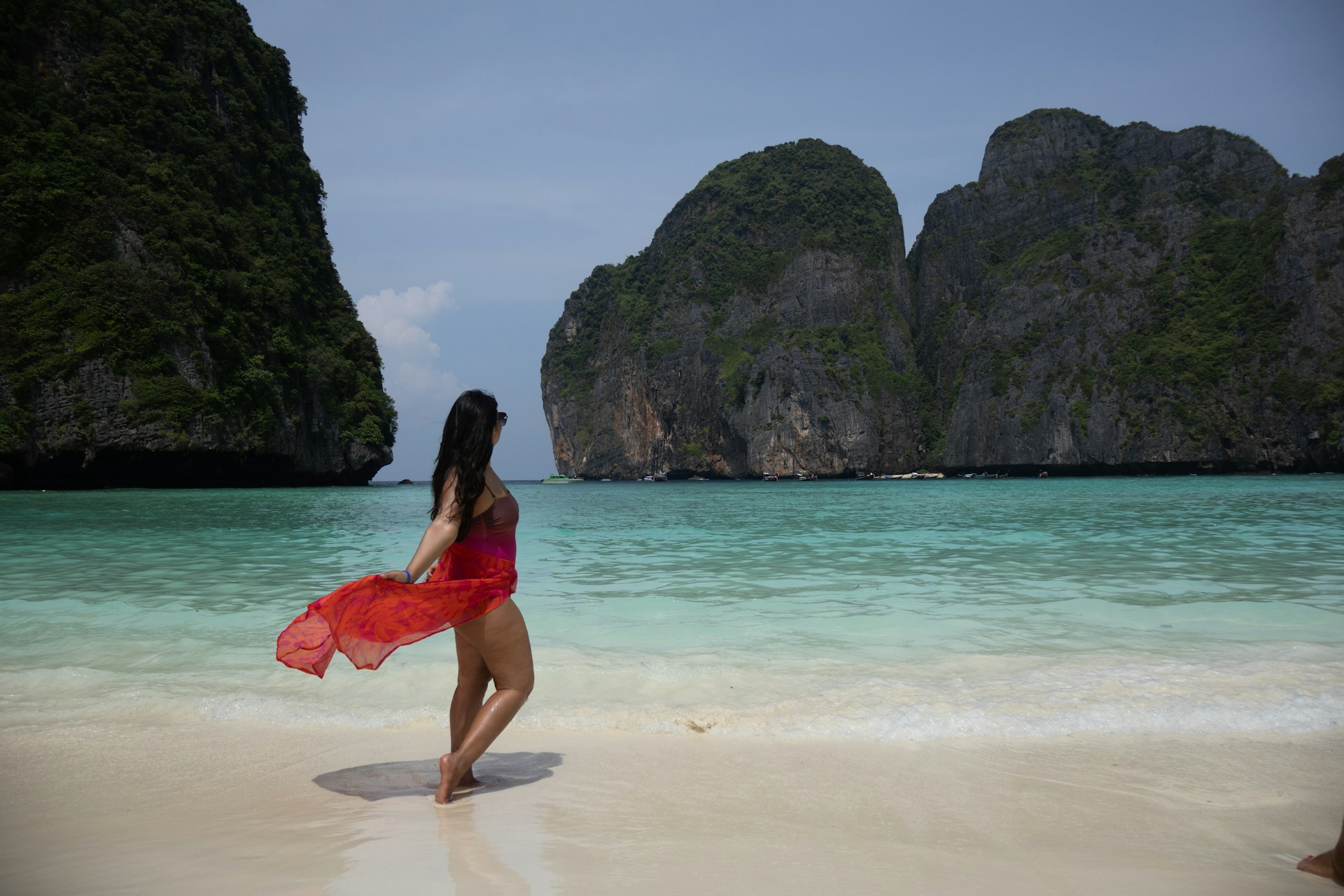 A woman wearing a flowing red dress stands on the sandy beach, gazing at the ocean under a bright blue sky.