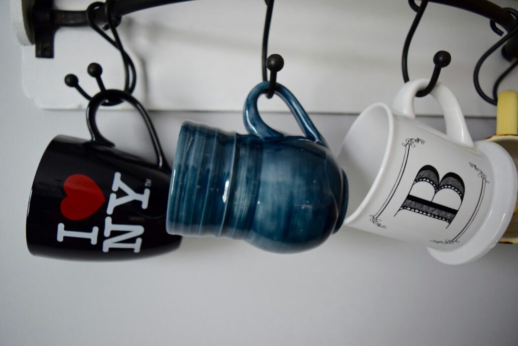 Three mugs -- one black with "I heart NY" written on it, one blue and one white with the letter "B" in black, hang on three black hooks on a white wall