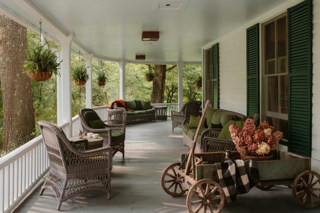 A wrap-around porch with hanging plants, three wicker chairs and two wicker couches with green cushions, and a decorative wooden wagon
