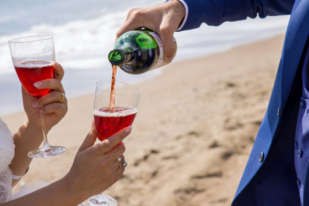 On a beach, a groom pouring wine from a bottle into a second glass held by a bride