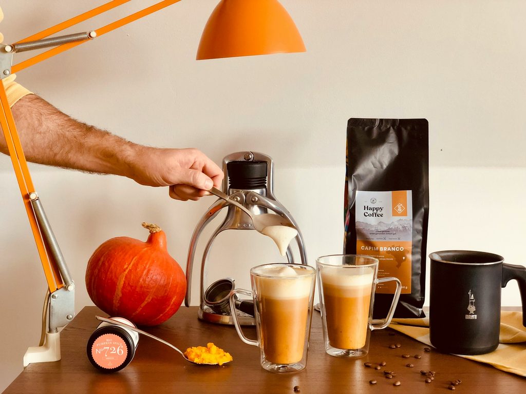 Arm stirring pumpkin latte on table with a coffee machine, coffee beans, spoon with pumpkin puree, orange pumpkin