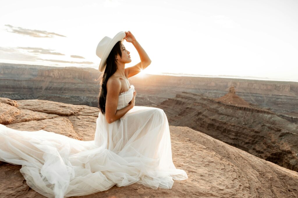 A bride wearing a white gown and cowboy hat posts in front of a canyon. 