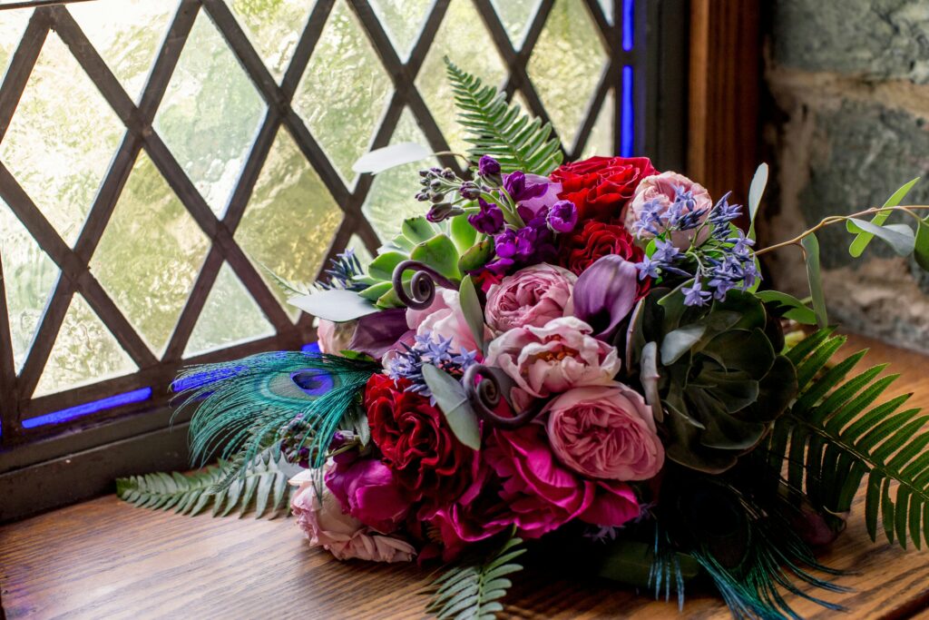 A bouquet of flowers featuring purple and pinks flowers, greenery and peacock feathers. 