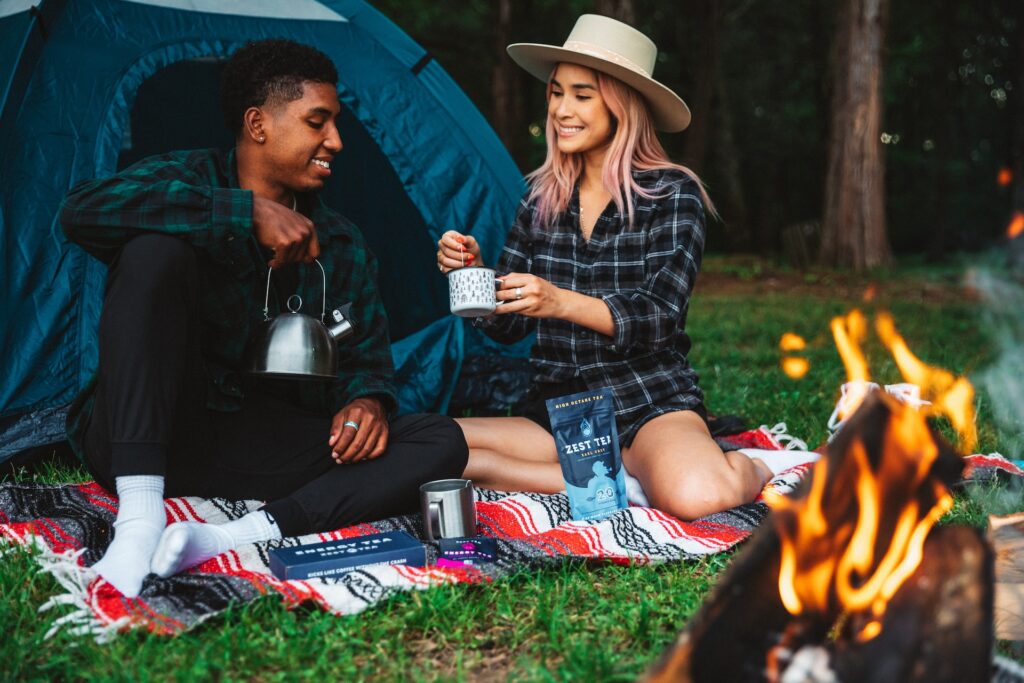 A man and woman drink tea by a tent and a campfire.