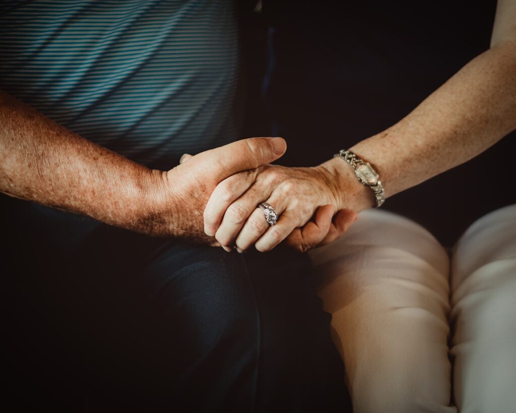 An older couple holds hands, with a wedding ring visible on the woman's finger.
