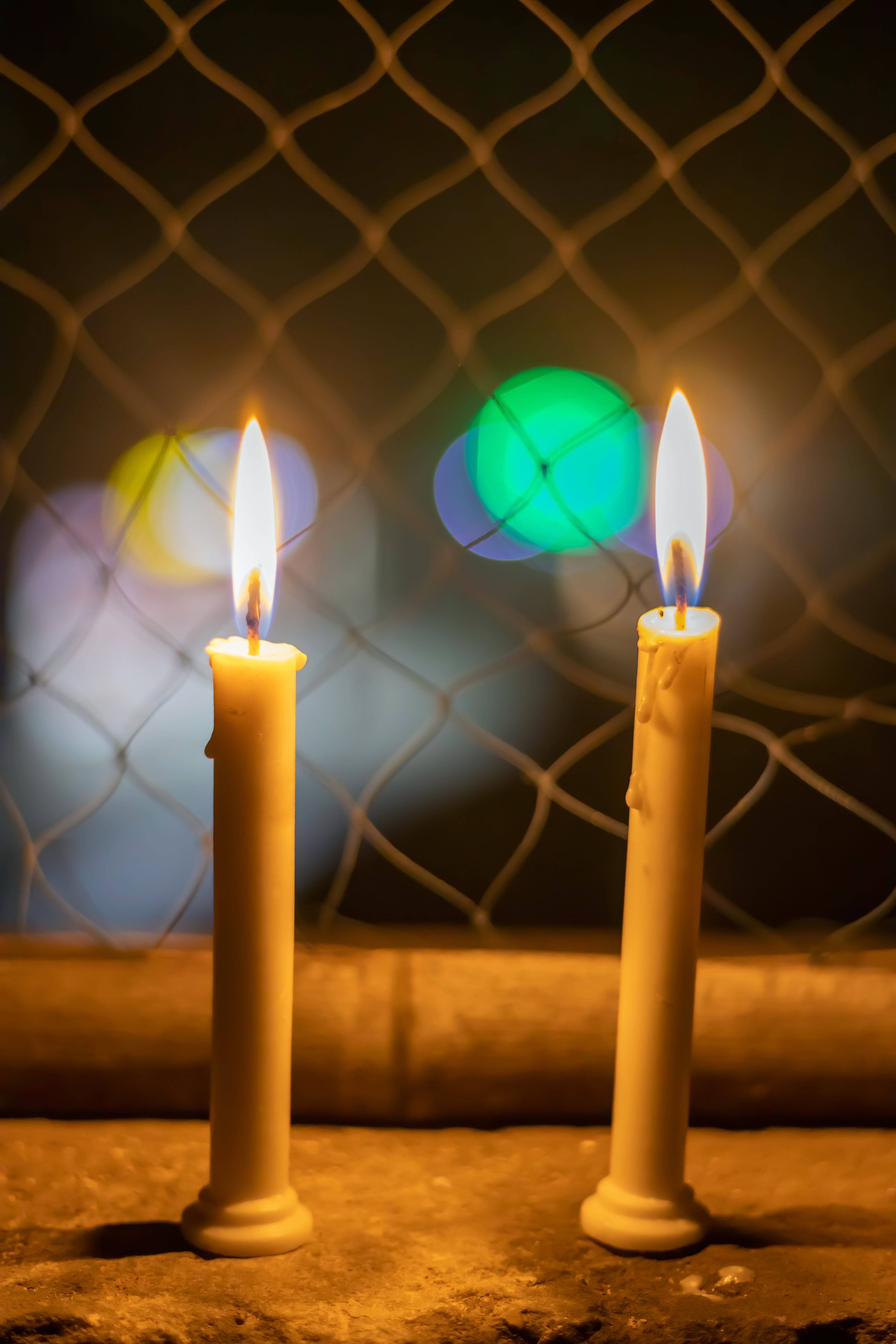 Two lit candles site on a ledge in front of a fence.