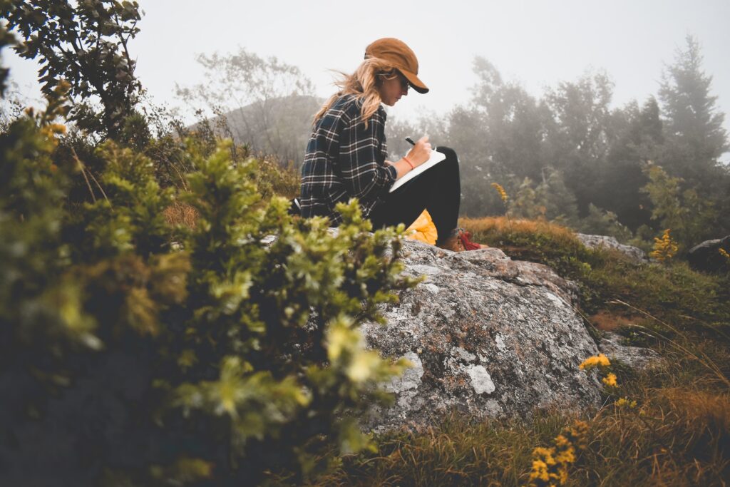 A woman sits on a rock in a field writing in a journal.
