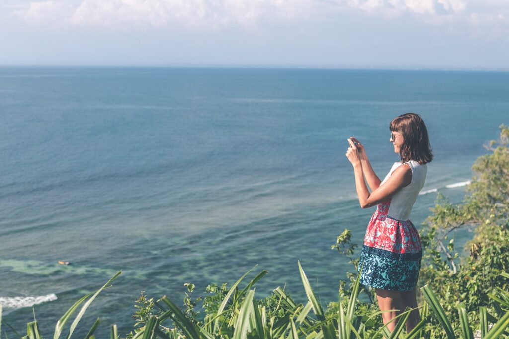 A woman in a blue, red and white dress takes a picture overlooking the ocean.